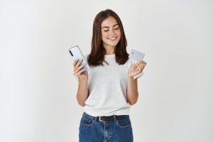 Portrait Happy Young Brunette Woman Showing Plastic Credit Card While Holding Mobile Phone Isolated White Wall Scaled