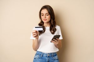 Image Young Happy Woman Paying Online Holding Smartphone Credit Card Enter Info Application Mobile Phone Standing Against Beige Background 1 Scaled